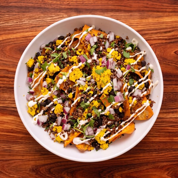 A white bowl of colorful nachos with sweet potato chips, black beans, quinoa, onions, and a creamy drizzle on a wooden table.