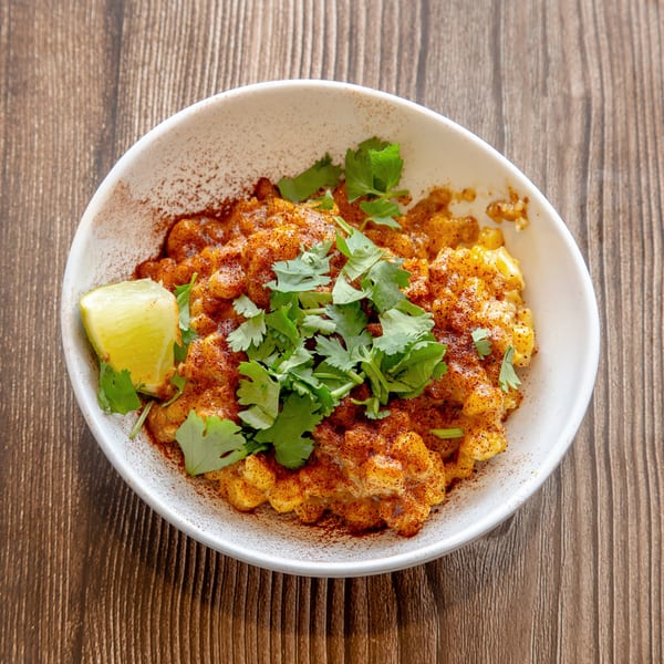 Elote-style corn in a white bowl, topped with cilantro, chili powder, and a lime wedge.