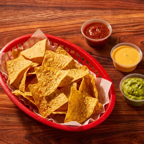 Tortilla chips in a red basket with salsa, cheese dip, and guacamole on a wooden table.