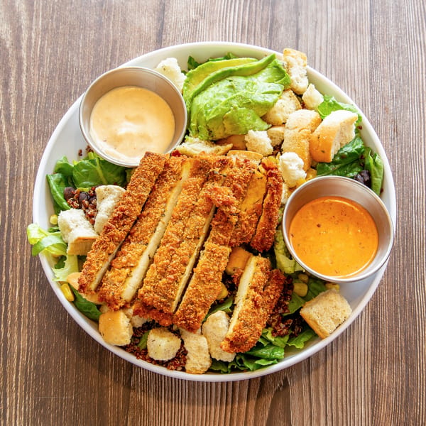 Crispy chicken salad with avocado, croutons, quinoa, and two dipping sauces on a wooden table.