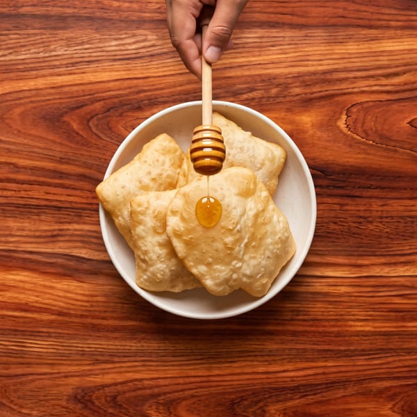 A hand drips golden honey from a dipper onto fried dough in a white bowl.