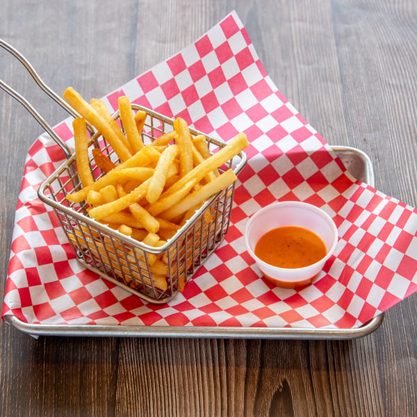 Golden french fries in a metal basket with a side of dipping sauce on red and white checkered paper.